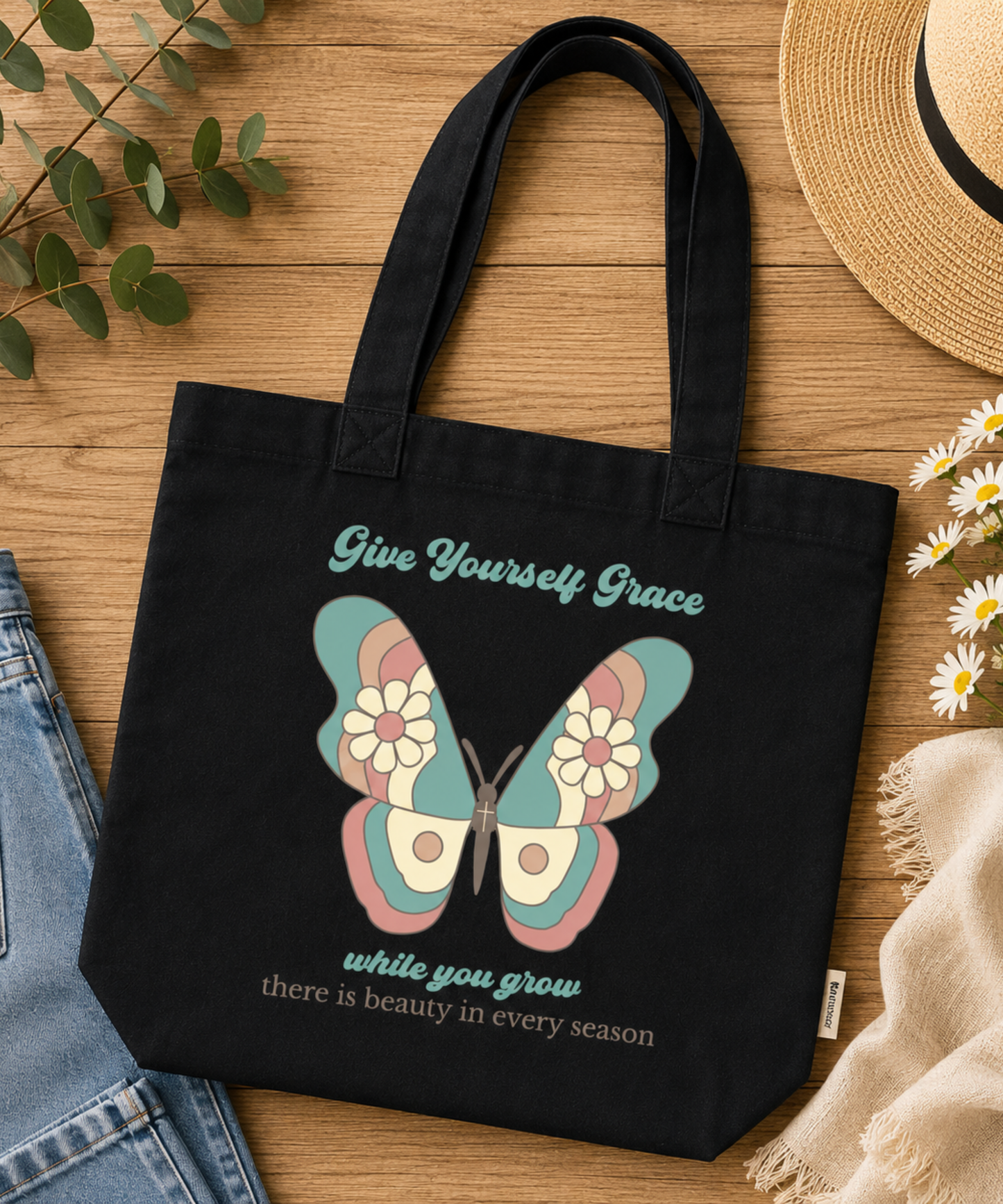 A black comfort colors canvas tote bag with a colorful butterfly graphic and the text 'Give Yourself Grace while you grow there is beauty in every season,' styled on a wooden surface with jeans, a straw hat, flowers, and a scarf.