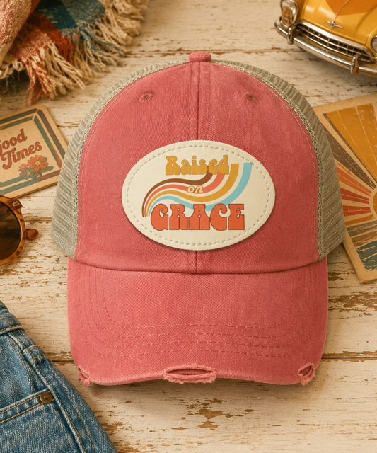 A distressed trucker cap in distressed coral and tan mesh, featuring an oval front patch that reads 'Raised by Grace' in a retro rainbow design. The hat is styled on a rustic wooden surface with jeans, sunglasses, cards, and a yellow car prop.
