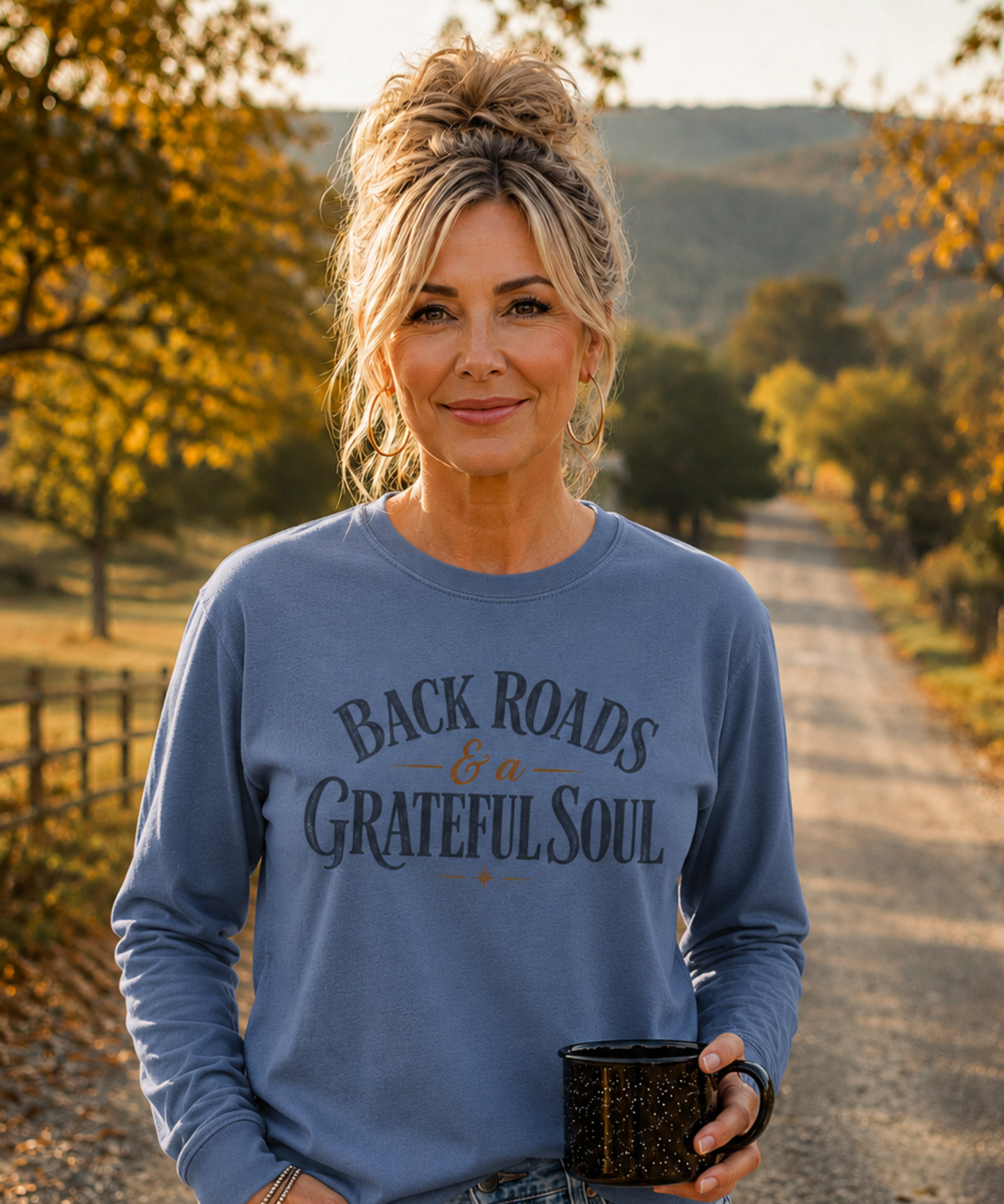 A woman stands on a gravel path outdoors wearing a blue jean comfort colors long-sleeve graphic shirt that reads 'Back Roads & a Grateful Soul' in dark lettering. She is holding a black speckled mug, with trees and hills in the background.