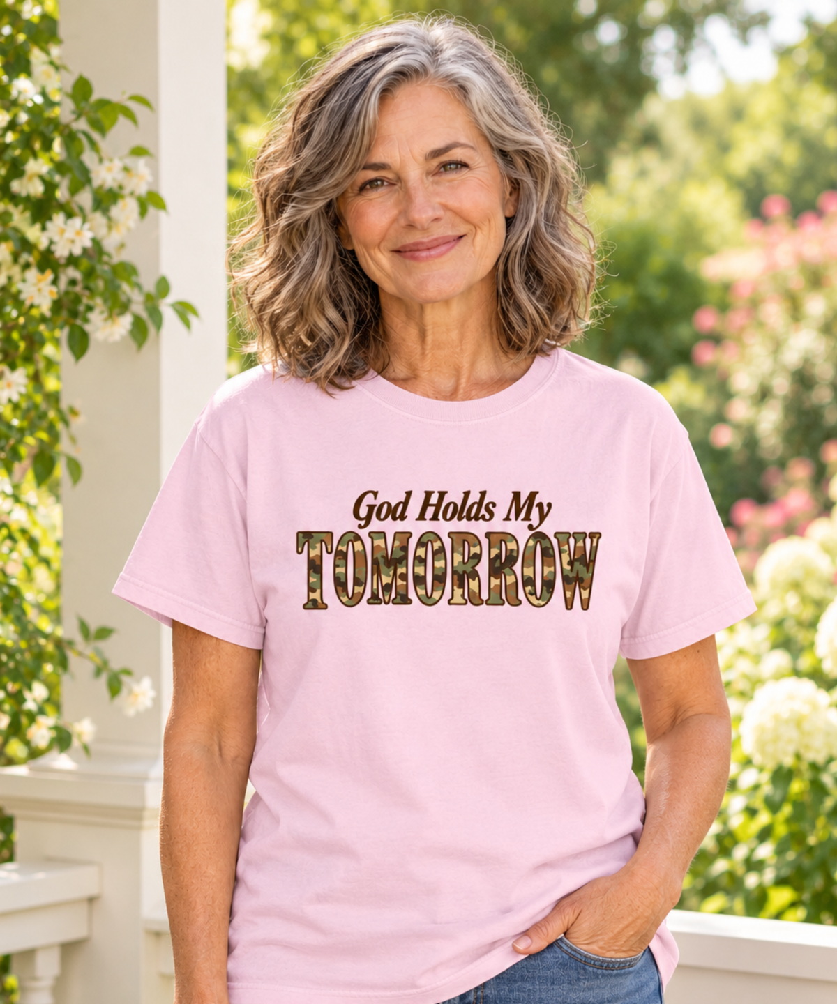 Woman with salt-and-pepper hair wearing a blossom pink God Holds My Tomorrow camo lettering tee and jeans on a cream porch overlooking a garden.