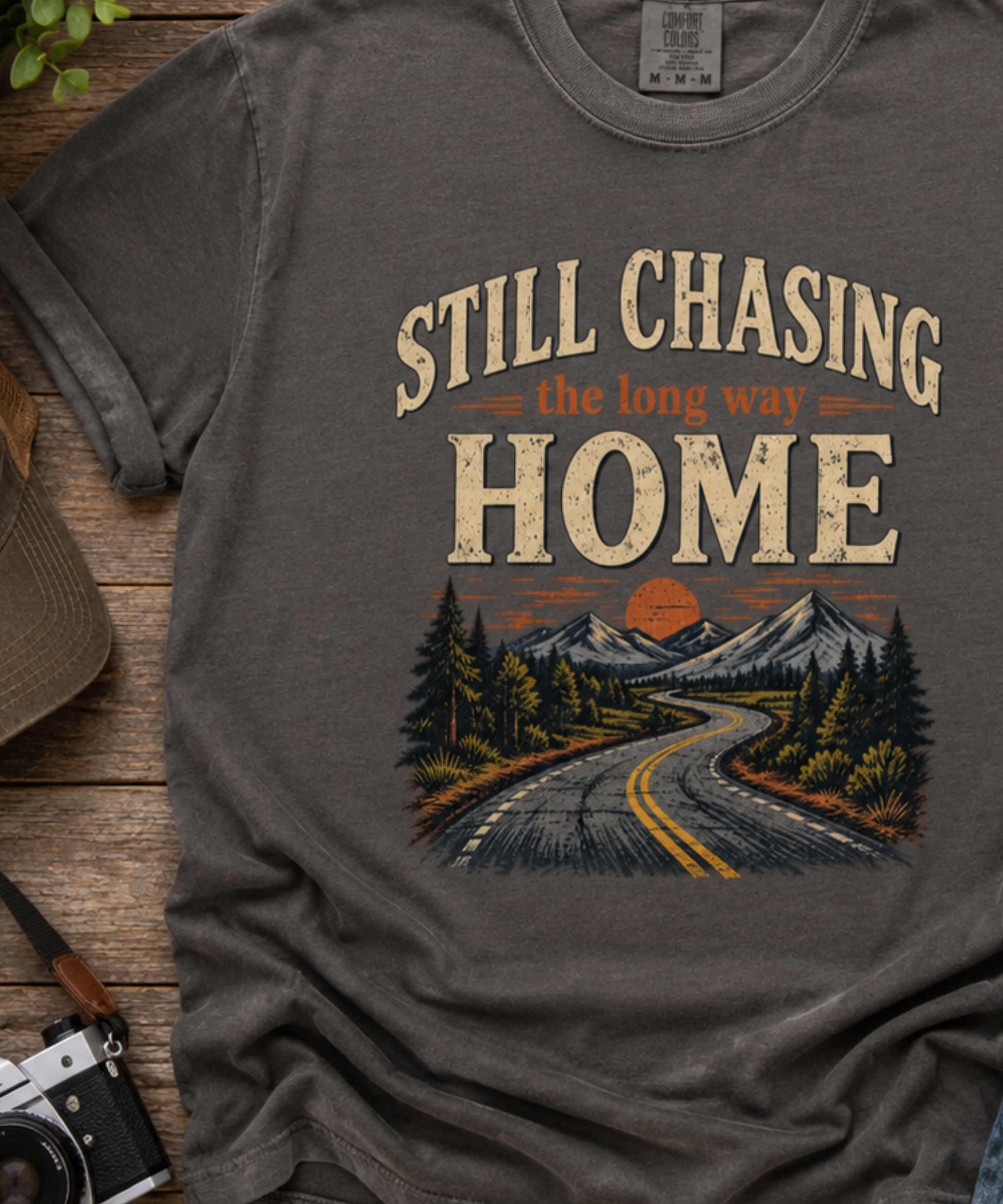 Close up of a pepper graphic t-shirt laid flat on a rustic wooden surface, featuring a vintage-style scenic print of a winding road through pine trees toward mountains at sunset, with the text 'STILL CHASING the long way HOME'.