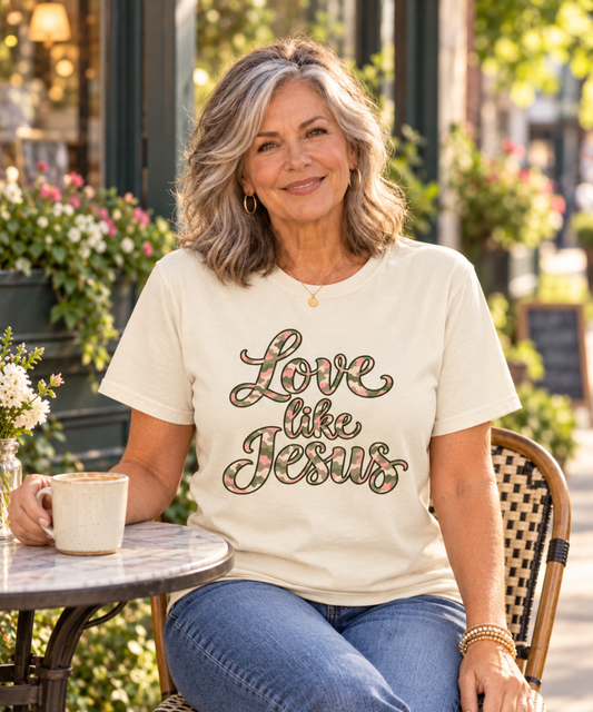 A woman sitting at an outdoor cafe table wearing an ivory comfort colors t-shirt with 'Love like Jesus' printed in a camo with pink, cream and green design. She is wearing blue jeans and holding a mug.