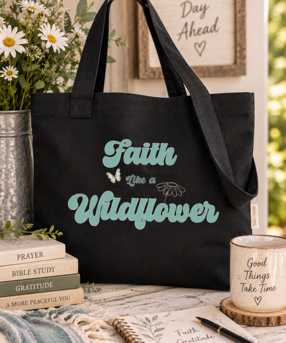 A black canvas tote bag with the phrase 'Faith is a Wildflower' printed in teal lettering, styled on a rustic wooden table with books, a mug, and flowers.