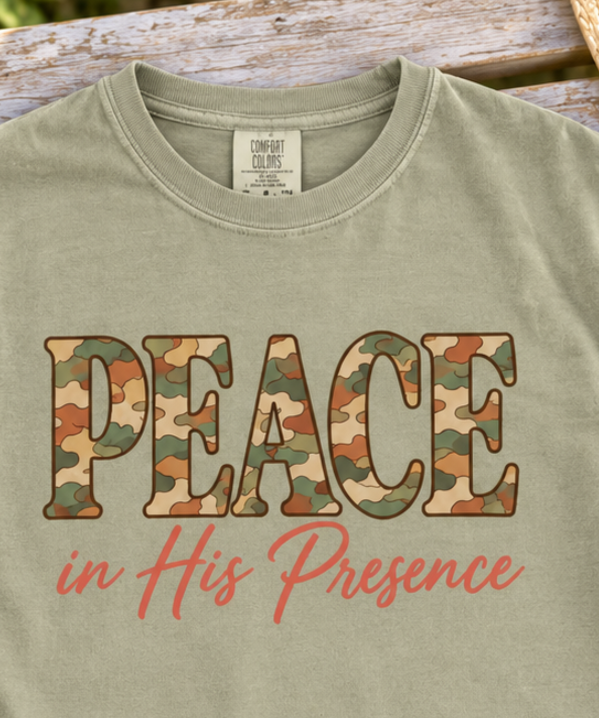 Close up of a khaki comfort colors t-shirt laid flat on a rustic wooden bench, featuring the text 'PEACE in His Presence' in camouflage and coral script. Decorative flowers and a straw hat frame the edges of the shot.
