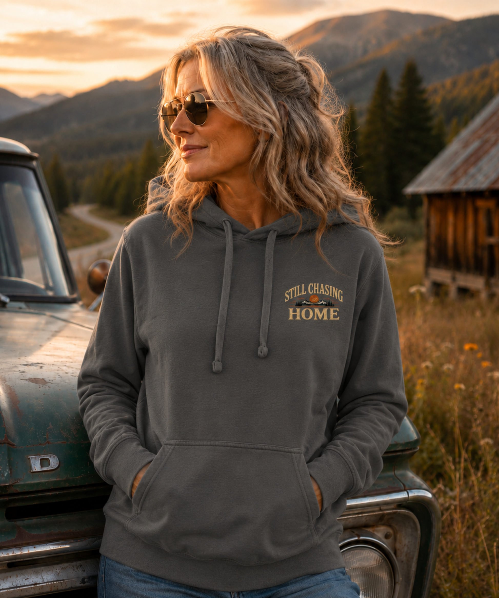 A woman wearing a pepper colored hoodie with 'Still Chasing Home' printed on the chest stands outdoors next to an old green truck, with mountains and a cabin in the background.