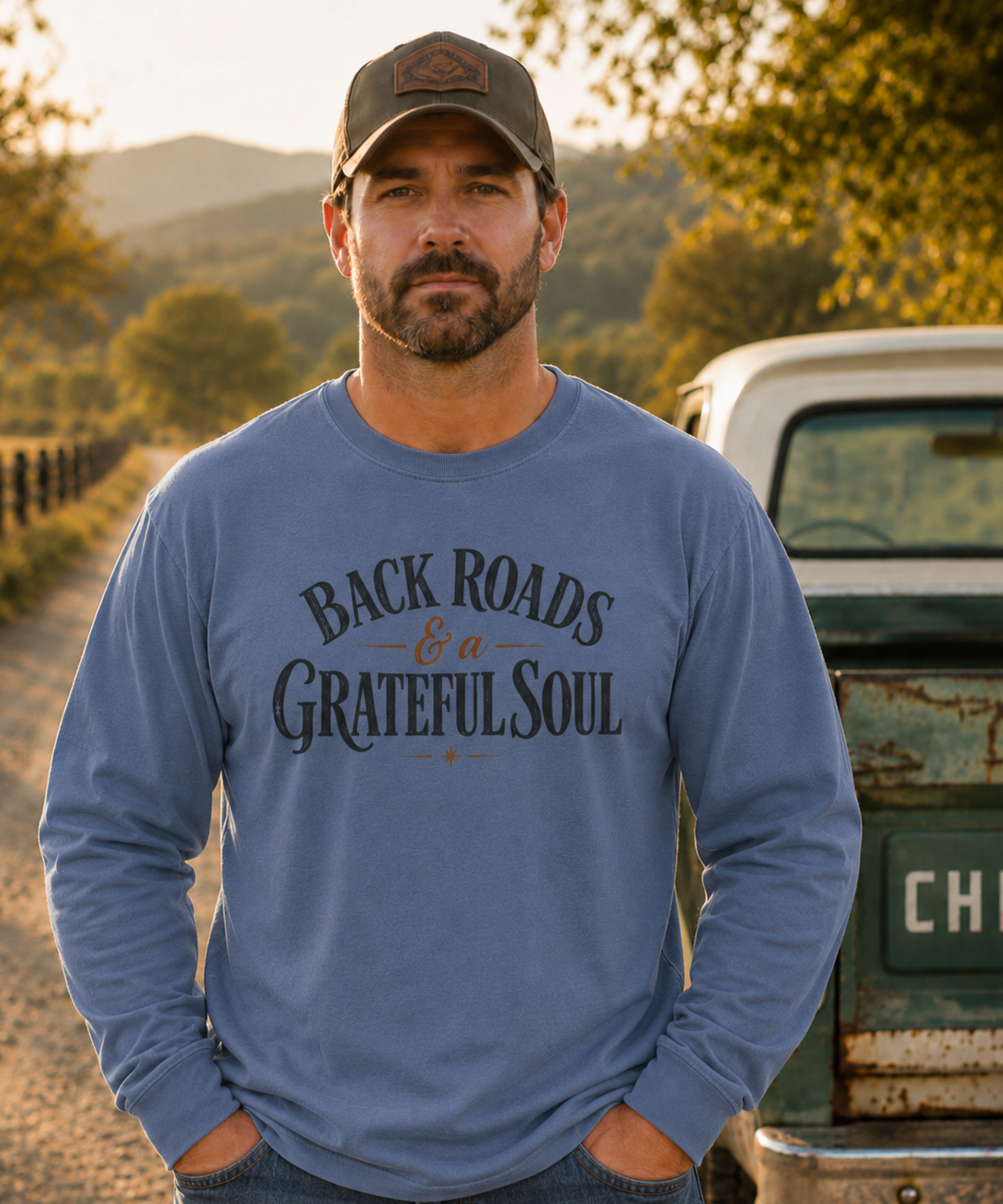 A man wearing a blue jean comfort colors long-sleeve t-shirt with 'BACKROADS & a GRATEFULSOUL' printed on the front, standing outdoors in front of a rustic white truck and a scenic background.