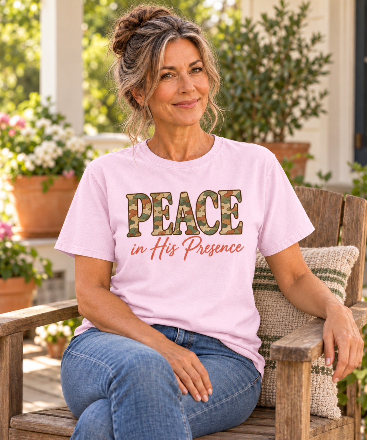 A woman sitting on a wooden porch chair wearing a light pink graphic t-shirt with the text 'PEACE in His Presence' in camouflage and coral script, paired with blue jeans.