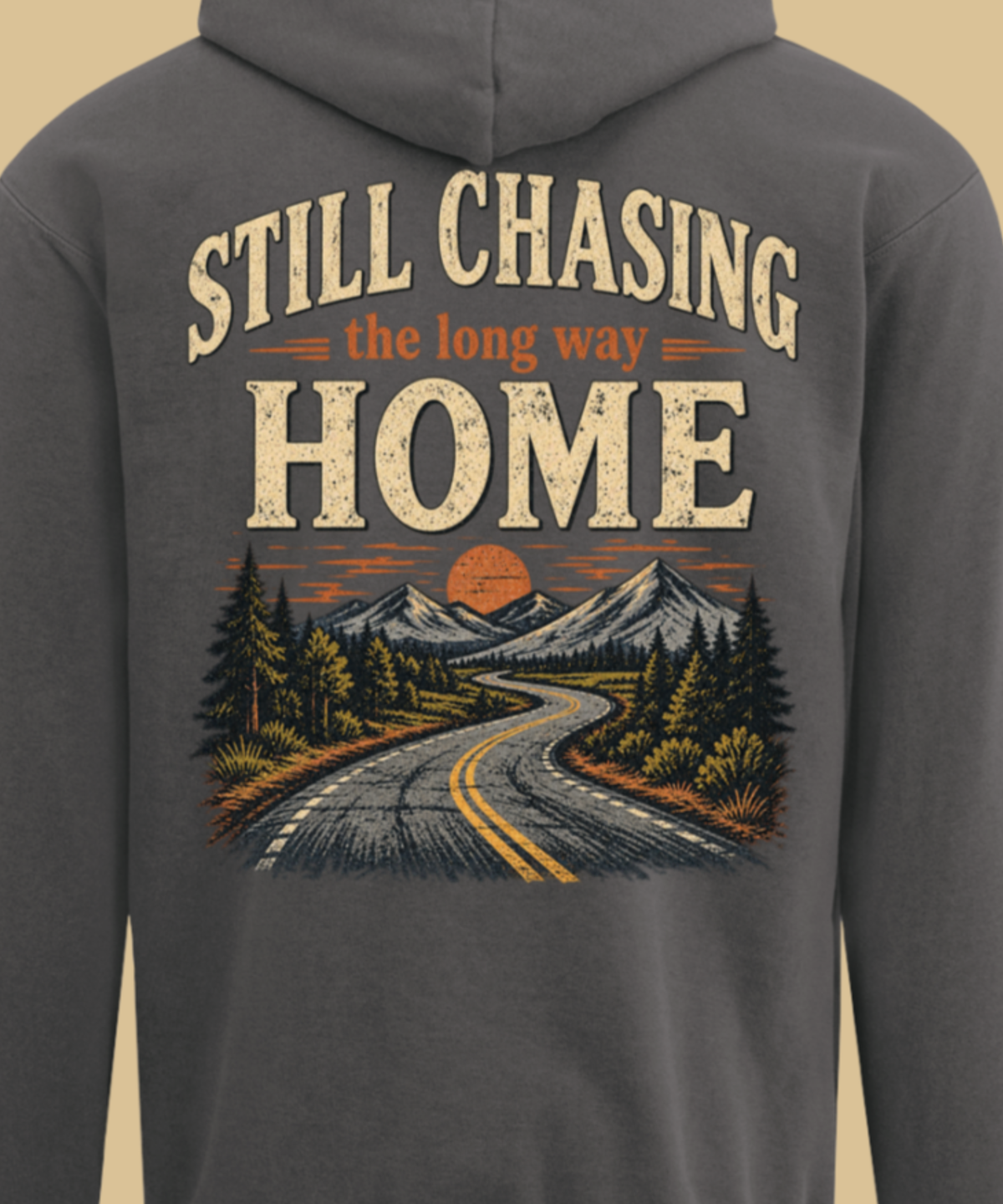 A pepper colored hooded sweatshirt shown from the back, featuring a large graphic print with the text 'STILL CHASING the long way HOME' above a scenic illustration of a winding road, mountains, trees, and a sunset.