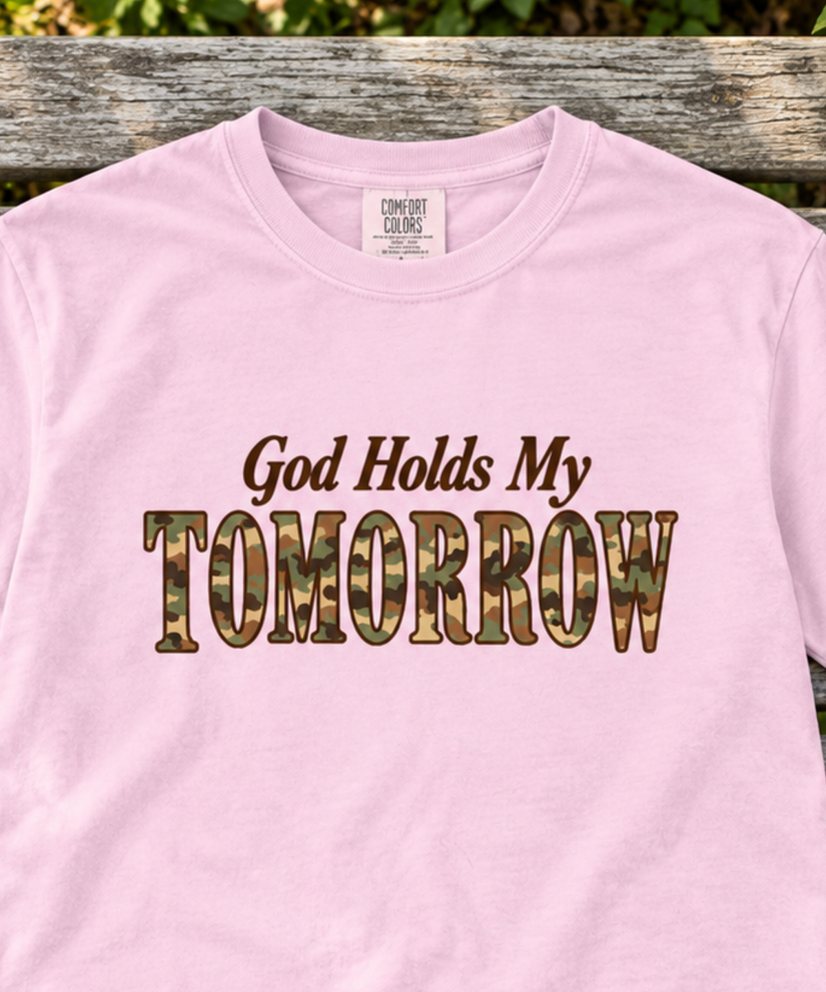 Blossom pink Comfort Colors tee featuring God Holds My Tomorrow in brown and camo lettering, laid flat on a weathered garden bench beside small white flowers.