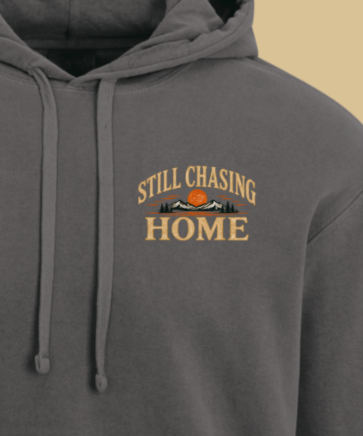 A close-up of a pepper colored hoodie featuring a graphic print on the chest that reads 'STILL CHASING HOME' in tan lettering, with a small orange sun and black mountain silhouette in the center.