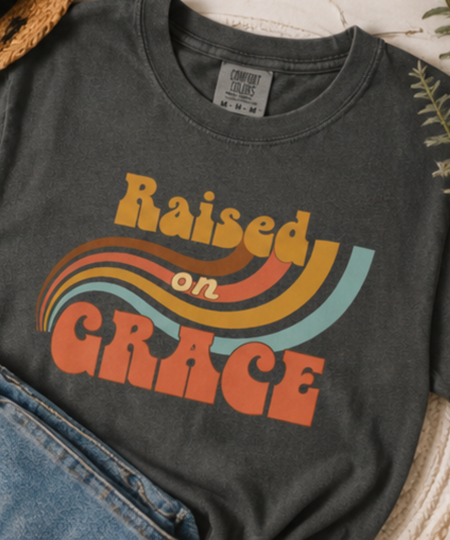 A graphite comfort colors t-shirt featuring a retro-style graphic that reads 'Raised on GRACE' in orange, yellow, and light blue wavy stripes. The shirt is styled flat with a pair of blue jeans and a woven straw hat visible around it.