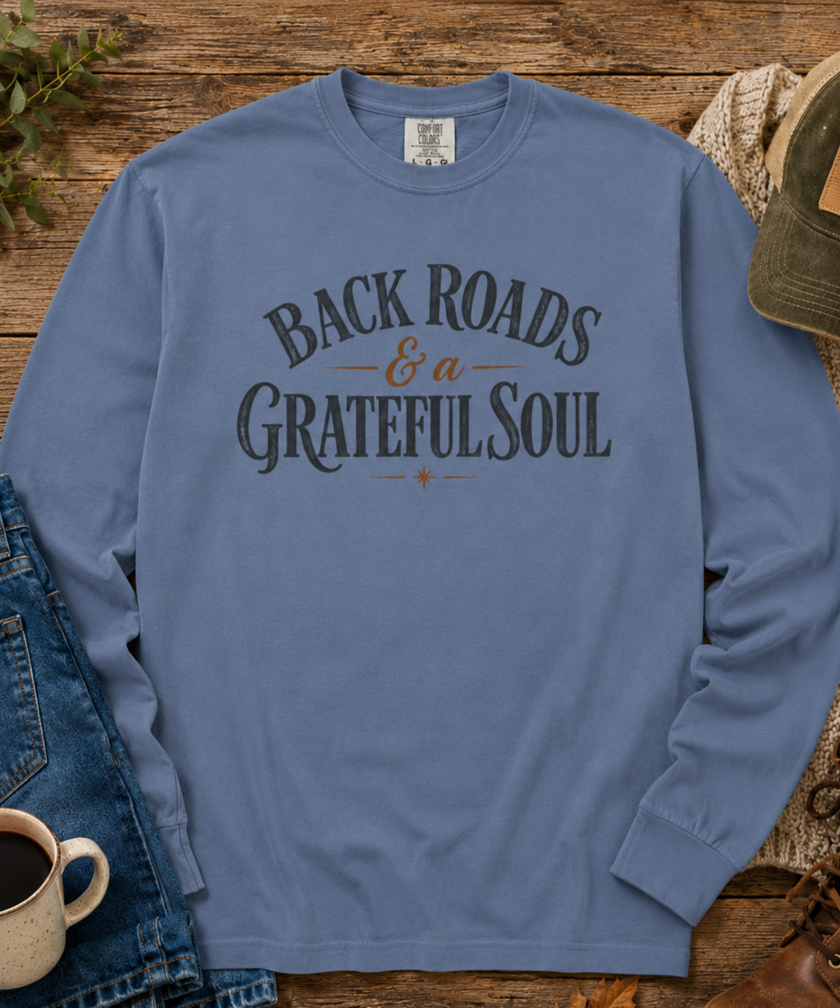 A blue jean comfort colors long-sleeve t-shirt with 'BACK ROADS & a GRATEFUL SOUL' printed in dark gray and brown on the chest, styled in a flat-lay on a rustic wooden background with jeans, a coffee cup, and a hat.