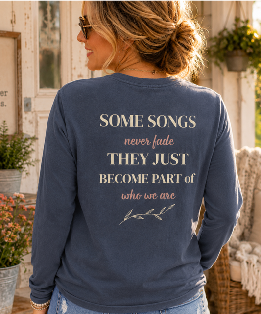 A woman shown from behind wearing a denim comfort colors long-sleeve t-shirt with a printed quote on the back that reads 'SOME SONGS never fade THEY JUST BECOME PART of who we are' in cream and dusty rose lettering, set against a rustic porch background.