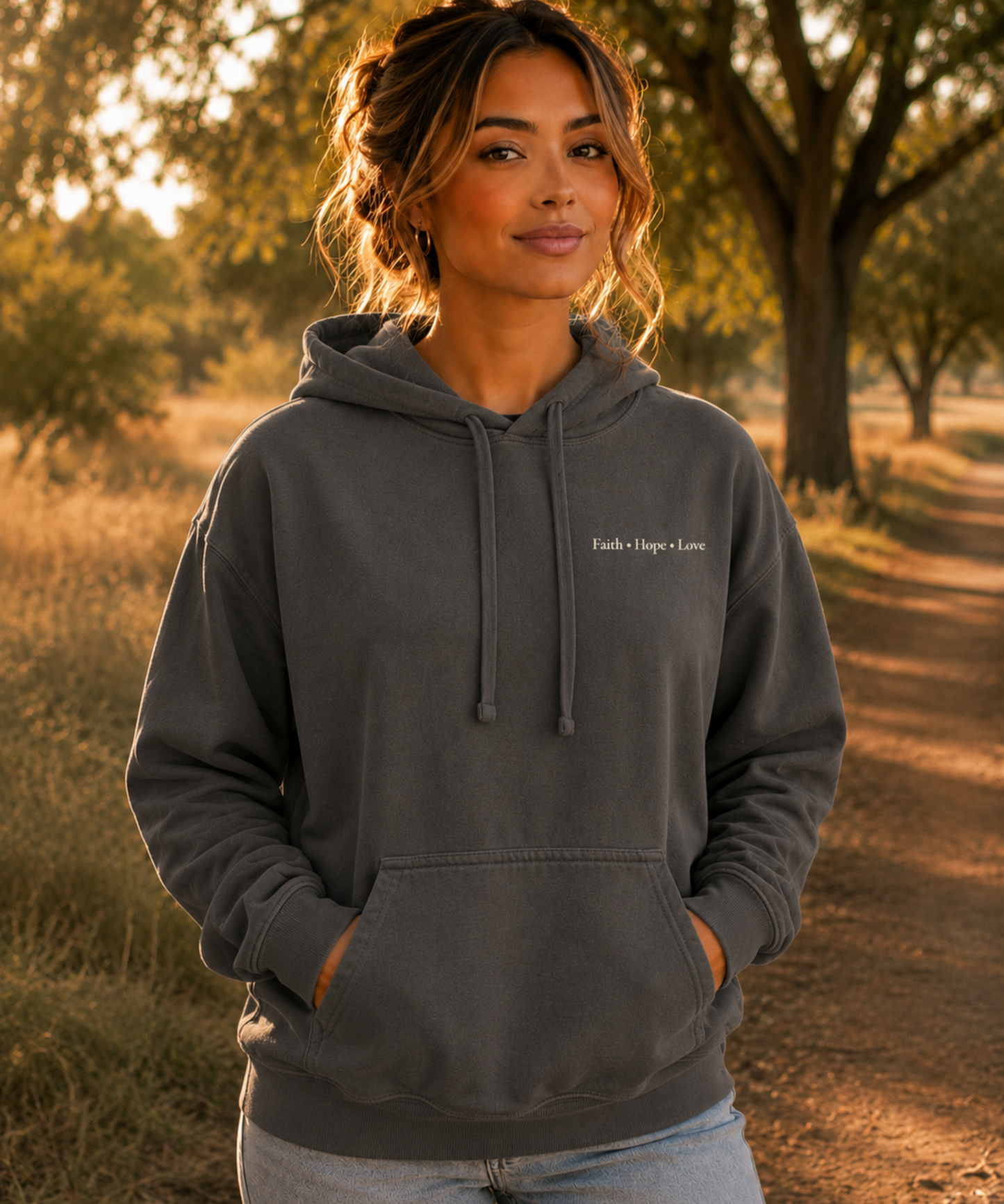 Woman wearing Comfort Colors pepper hoodie with Faith Hope Love left chest Christian design in a warm outdoor park