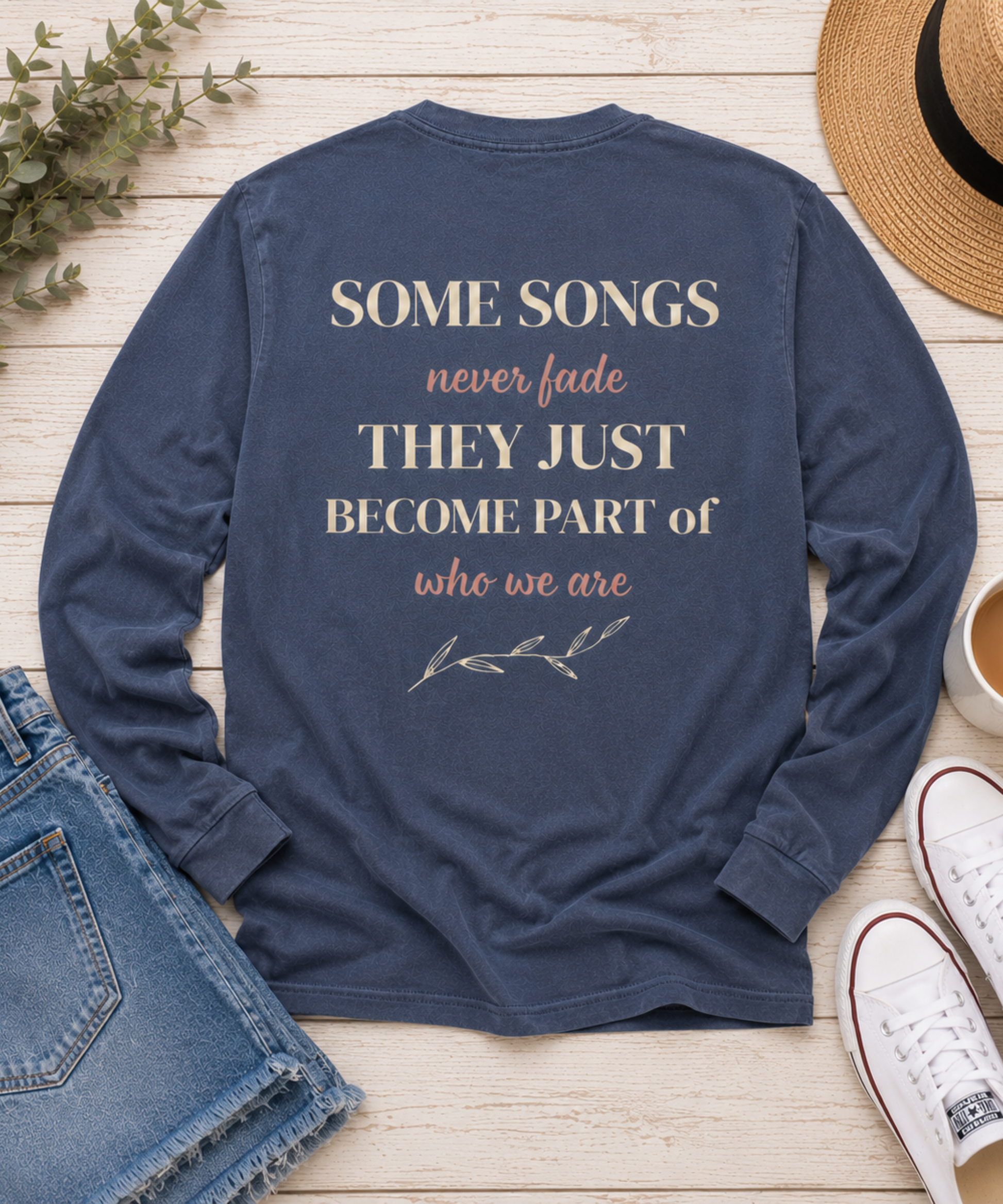 A denim comfort colors long-sleeve t-shirt with a printed quote on the back, laid flat on a light wood background with jeans, a straw hat, white sneakers, and a small plant sprig.