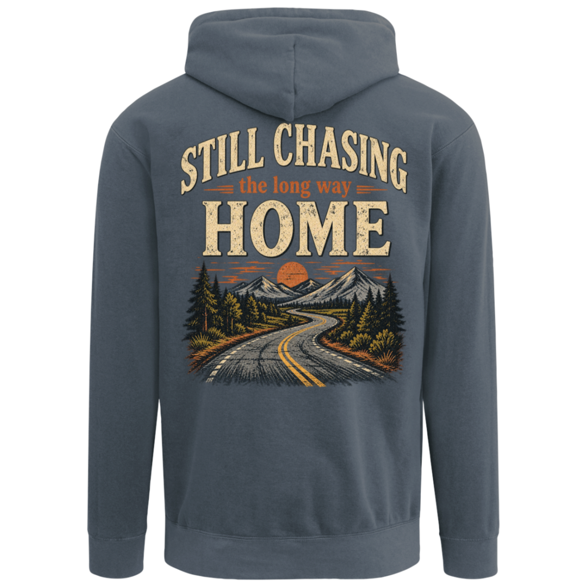 A denim comfort colors hoodie shown from the back, featuring a large graphic print with the text 'STILL CHASING the long way HOME' above an illustration of a winding road, pine trees, mountains, and an orange sunset. Includes left chest design.