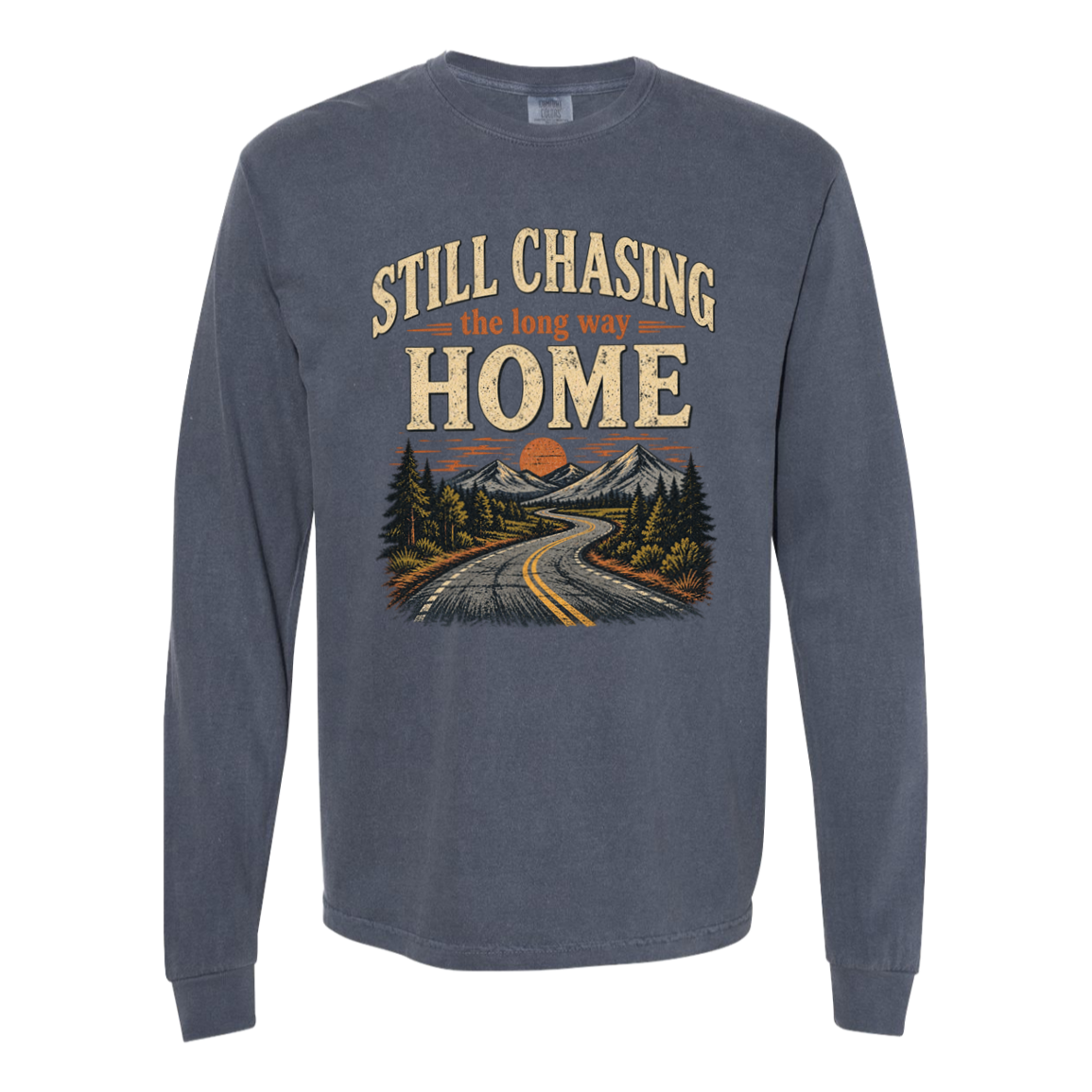 A long-sleeve crewneck t-shirt in denim , featuring a vintage-style graphic on the front with the text 'STILL CHASING the long way HOME' above an illustration of a winding road through mountains and pine trees.