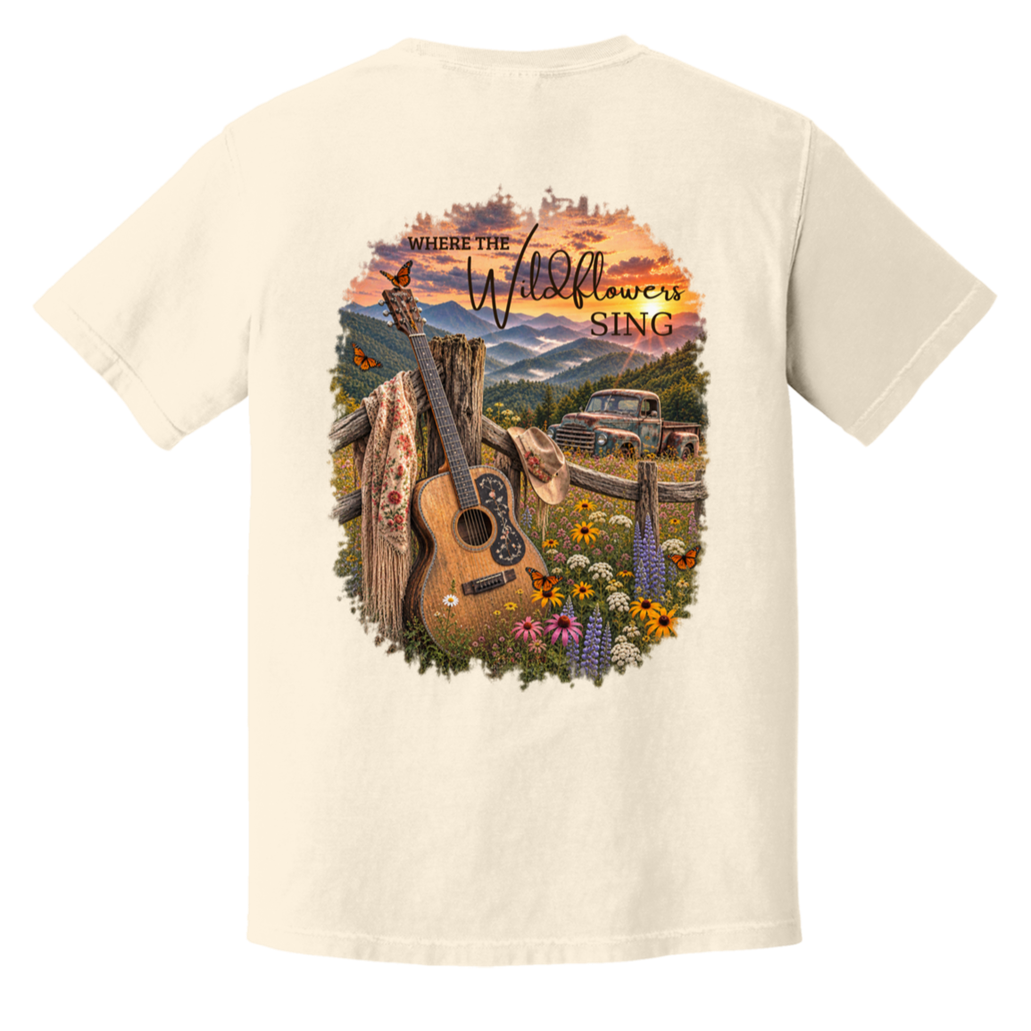 An ivory short-sleeve t-shirt showing the back, which features a large, colorful graphic print of an acoustic guitar, wildflowers, a rustic truck, and mountains under a sunset sky with the text 'WHERE THE Wildflowers SING'.