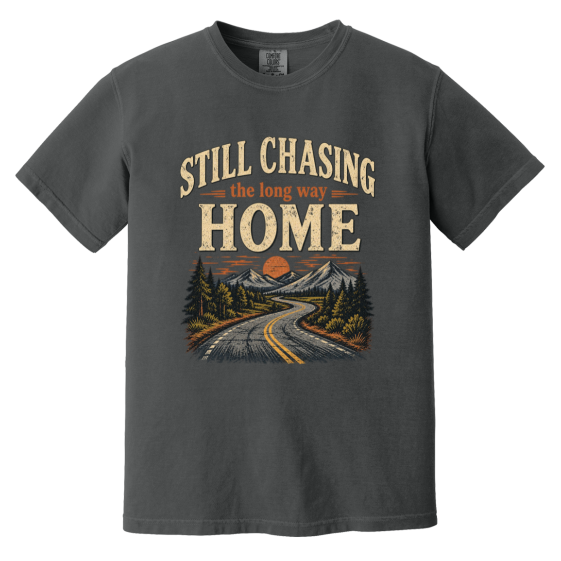 A graphite crewneck t-shirt featuring a vintage-style graphic print on the front. The graphic displays the text 'STILL CHASING the long way HOME' in distressed cream and orange lettering above an illustration of a winding road, pine trees, and mountains under an orange sunset.