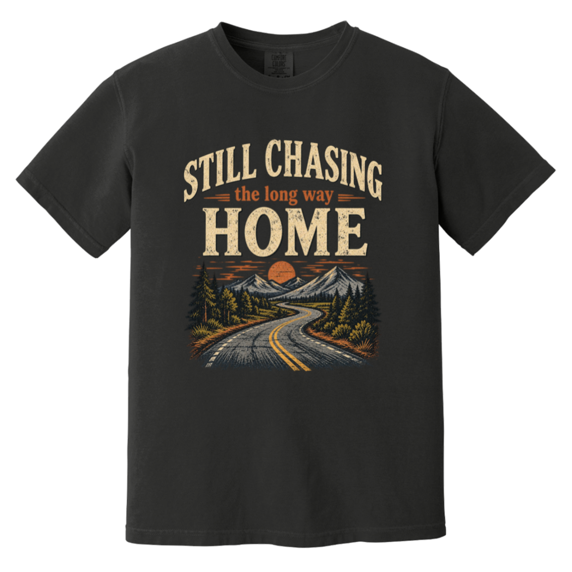 A black short-sleeve t-shirt with a vintage-style graphic on the front. The graphic features the text 'STILL CHASING the long way HOME' in distressed cream and orange lettering above an illustration of a winding road leading through pine trees and mountains toward a mountain peak under an orange sun.