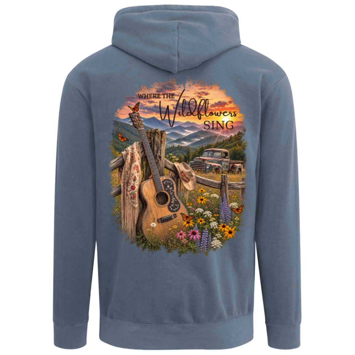 A blue jean comfort colors hoodie shown from the back, featuring a large, colorful graphic print of an acoustic guitar, wildflowers, a rustic fence, a vintage pickup truck, and mountains under a sunset sky with the text 'Where the Wildflowers Sing'. This has a left chest design of a butterfly.