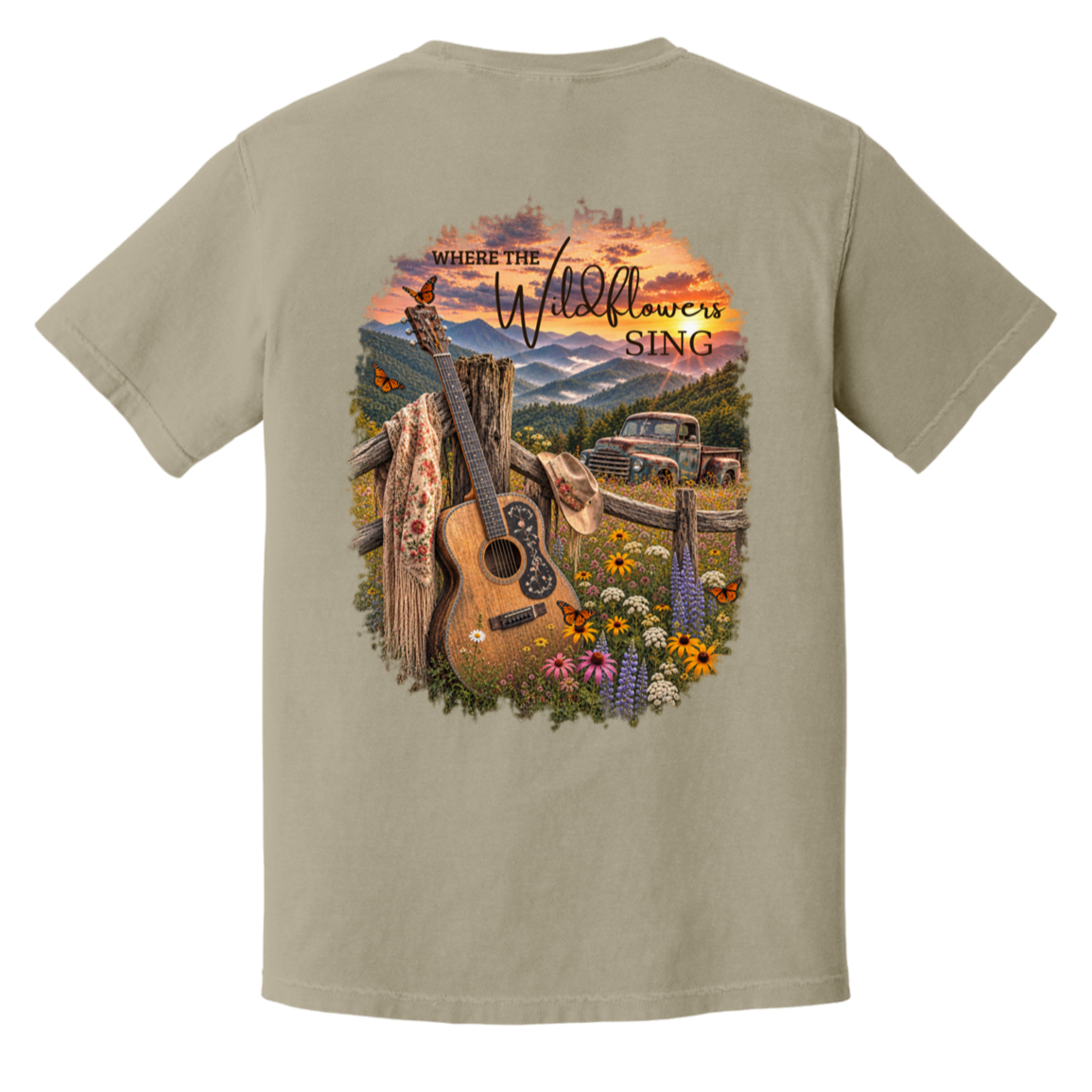 A sandstone crewneck t-shirt shown from the back, featuring a large, colorful graphic print of an acoustic guitar, wildflowers, a rustic truck, and mountains under a sunset sky with the text 'WHERE THE Wildflowers SING'.