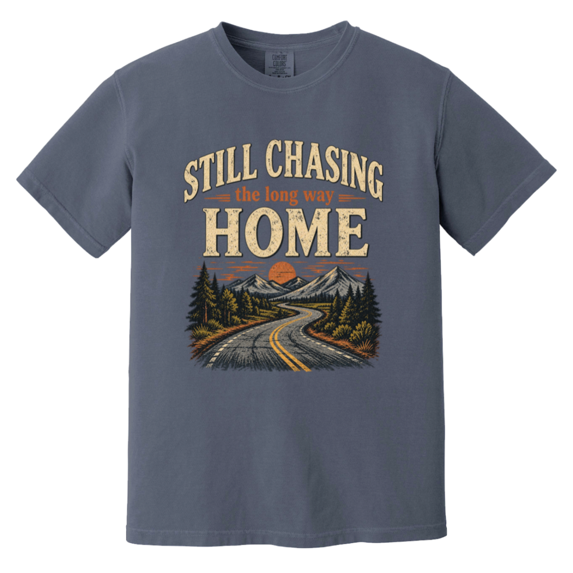 A denim crewneck t-shirt featuring a retro-style graphic print of a winding road leading through mountains and pine trees, with the text 'STILL CHASING the long way HOME'.