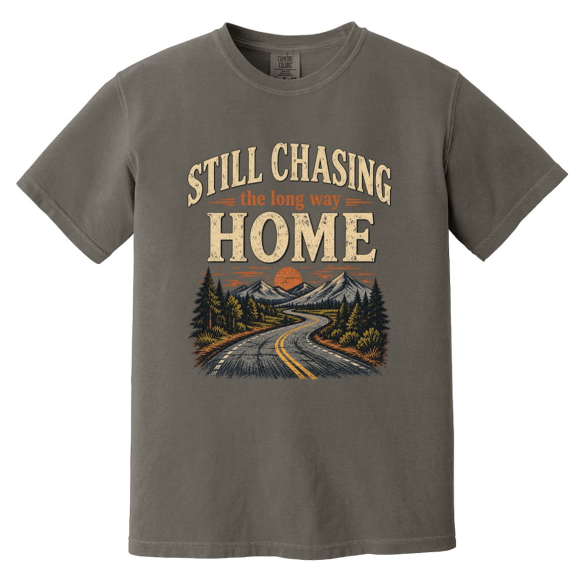 A short-sleeve crewneck t-shirt in a pepper color, featuring a retro-style graphic print on the front with the text 'STILL CHASING the long way HOME' above an illustration of a winding road through a mountain landscape.