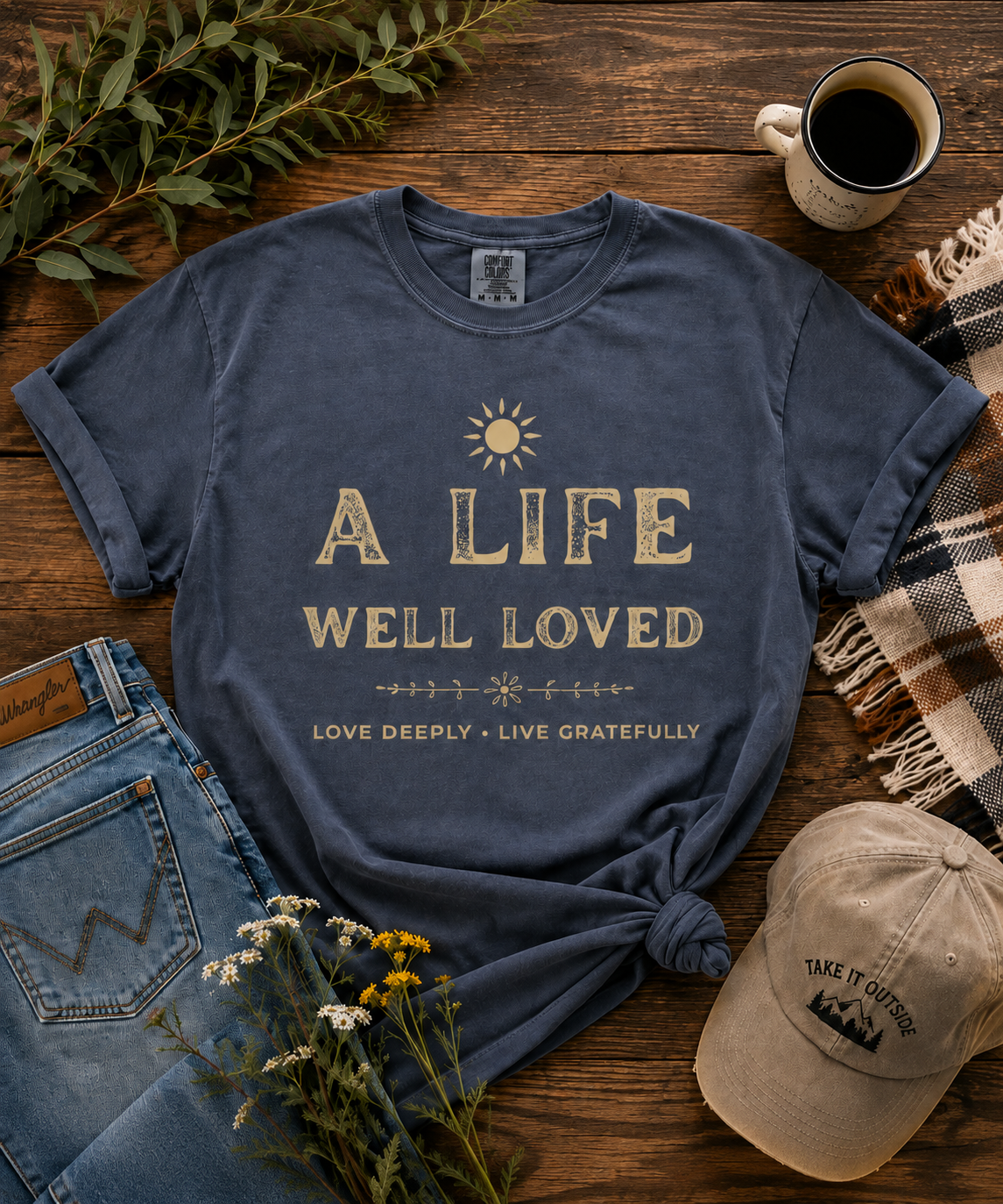 Vintage-inspired A Life Well Loved graphic T-shirt in faded denim blue with distressed cream lettering, sun design, and Love Deeply Live Gratefully message in a rustic outdoor flat lay.