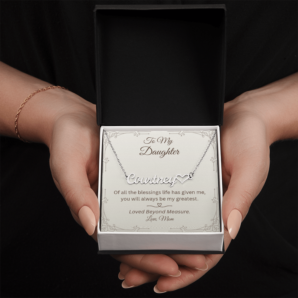 A silver nameplate necklace with a small heart pendant is displayed inside an open black gift box held by two hands. The necklace is mounted on a printed card addressed 'To My Daughter' with a personalized name and a sentimental message.