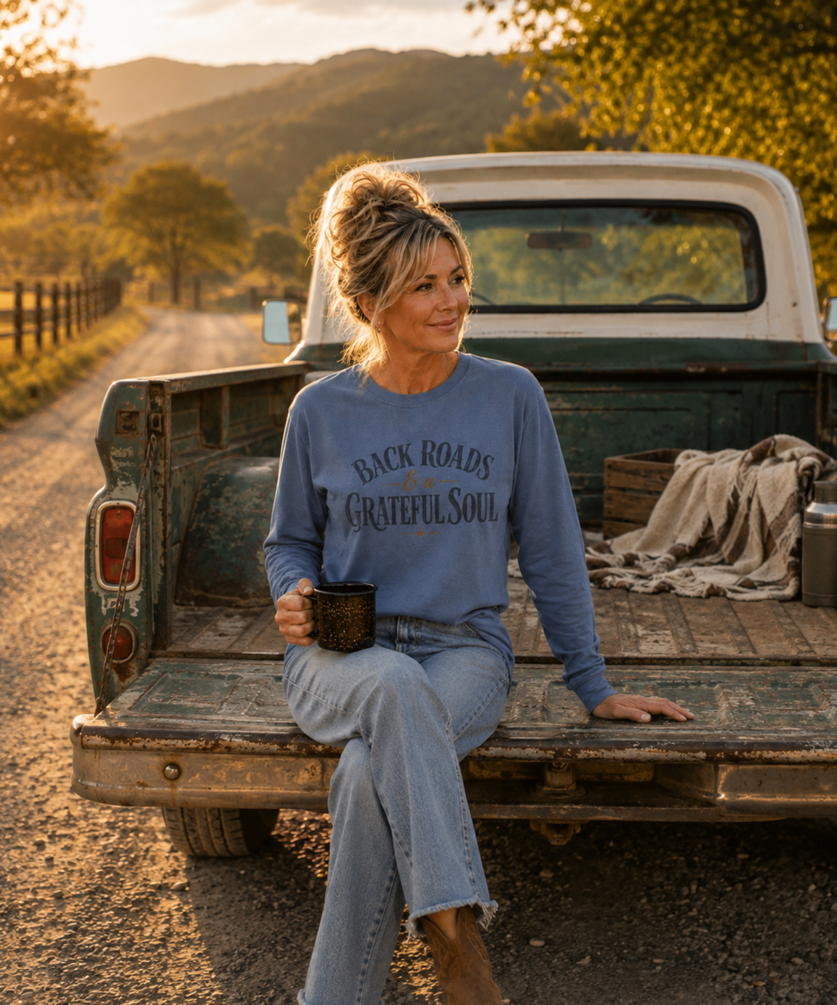 A woman sits on the tailgate of a vintage pickup truck on a sunlit country road, wearing a blue jean comfort colors long sleeve t-shirt, with the text 'BACK ROADS ARE GRATEFUL SOUL' and light-wash jeans.