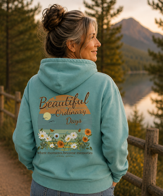 A woman wearing a seafoam hoodie with a floral graphic and the text 'Beautiful Ordinary Days' on the back, standing outdoors by a wooden fence with a mountain and lake in the background.