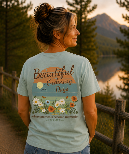 A woman wearing a bay comfort colors  graphic t-shirt with a sunset and wildflower design on the back, standing outdoors in front of a mountain and lake at sunset.