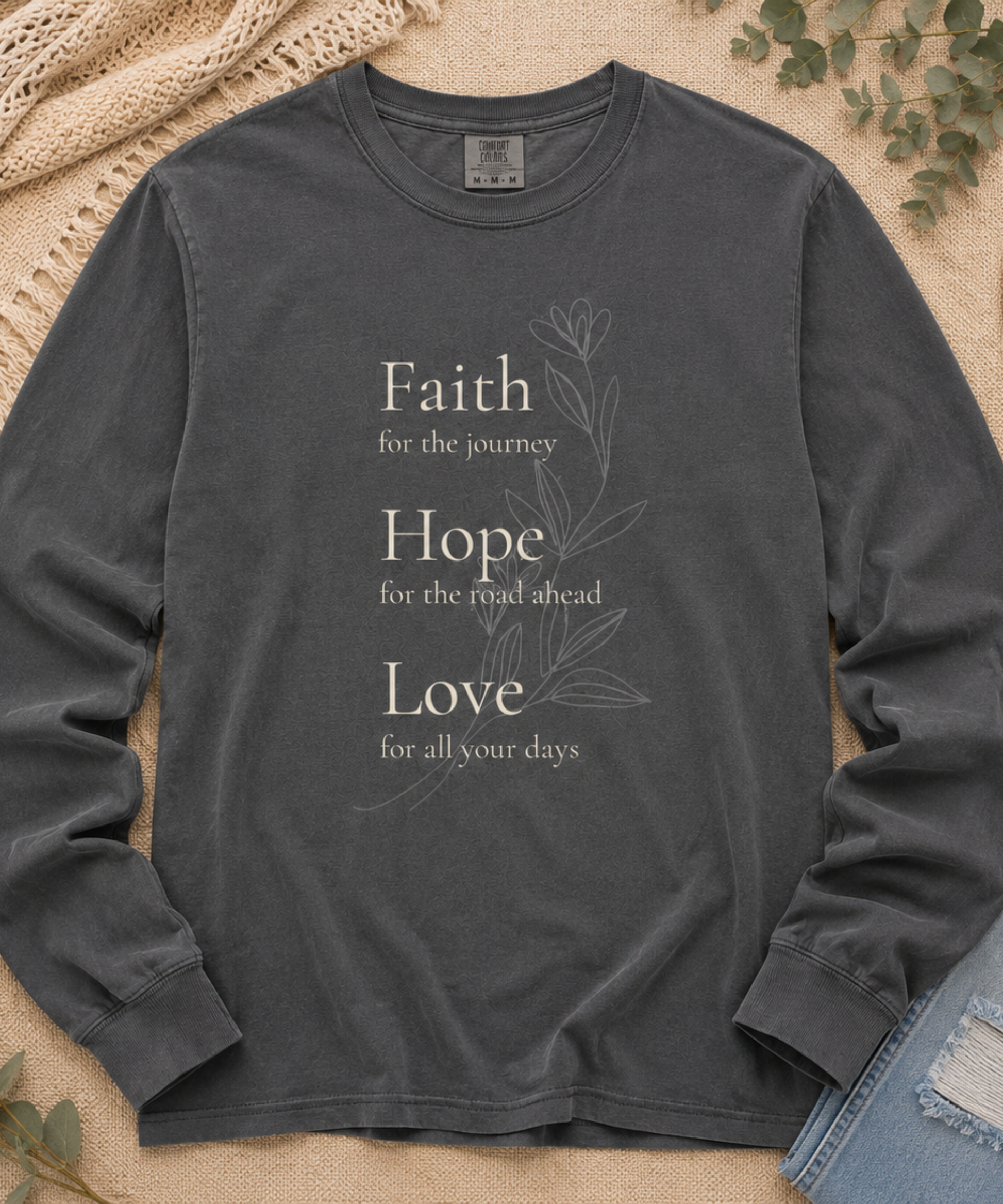 Flat lay of Comfort Colors pepper long sleeve shirt with Faith Hope Love botanical Christian front design on a neutral textured background.