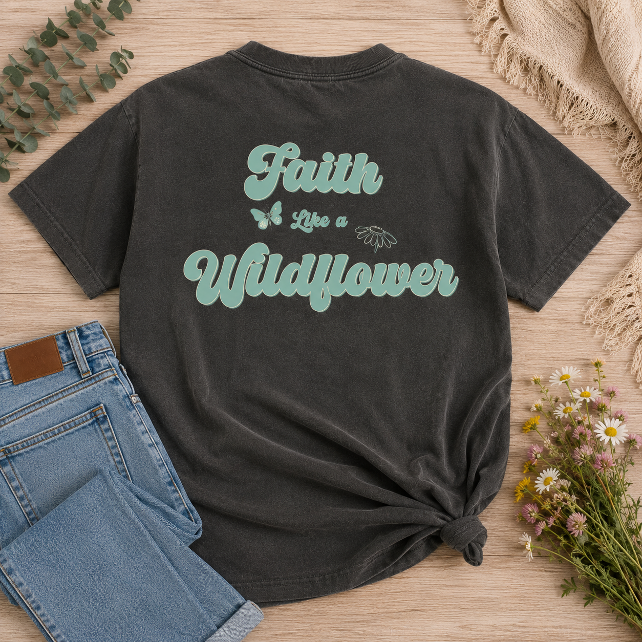 A Pepper comfort colors t-shirt with the phrase 'Faith Like a Wildflower' printed in a light teel green script, styled in a flat-lay on a light wood background with blue jeans, eucalyptus, and flowers.