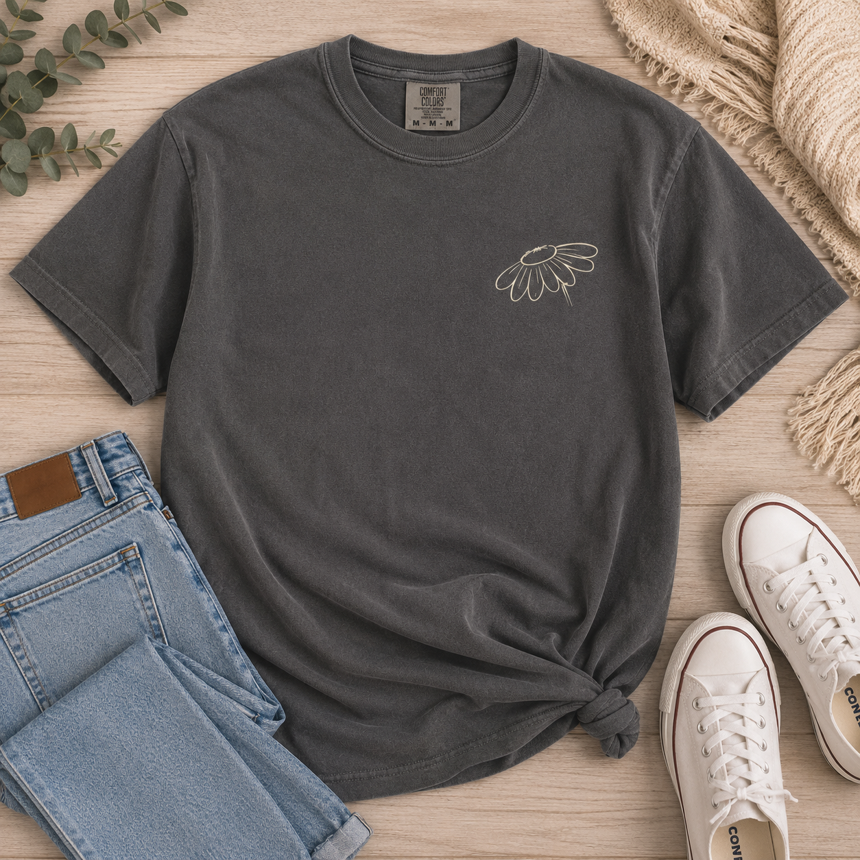 A Pepper comfort colors t-shirt with the phrase 'Faith Like a Wildflower' printed in a light teel green script, styled in a flat-lay on a light wood background with blue jeans, eucalyptus, and flowers.