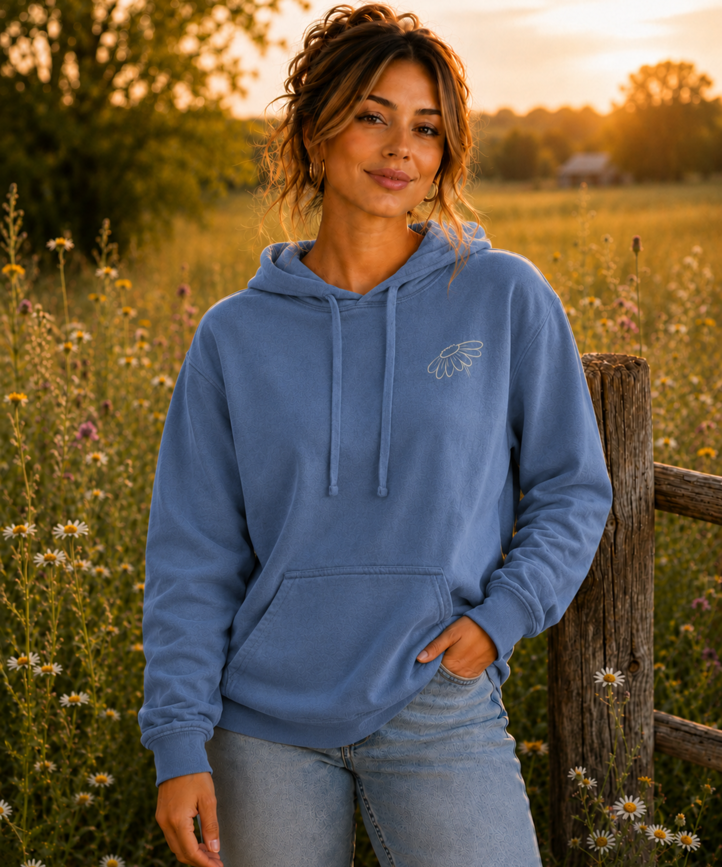 A woman wearing a Blue Jean comfort colors pullover hoodie with a small cream embroidered wildflower logo on the left chest, standing outdoors in a sunlit field of wildflowers next to a wooden fence.