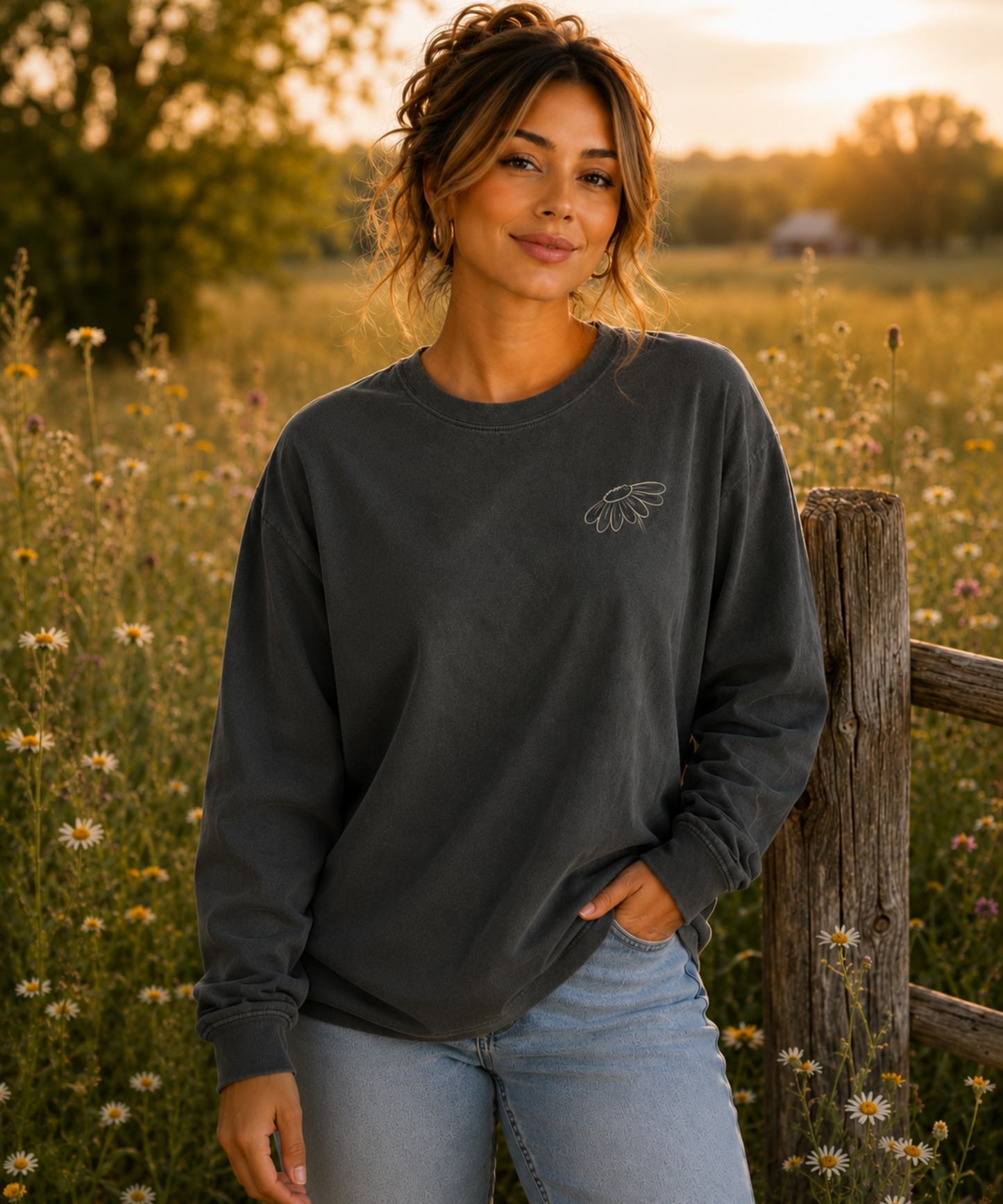 A woman stands in a sunlit field of wildflowers wearing a Pepper long-sleeve crewneck comfort colors shirt, with a small cream embroidered wildflower on the left chest, paired with light blue jeans.