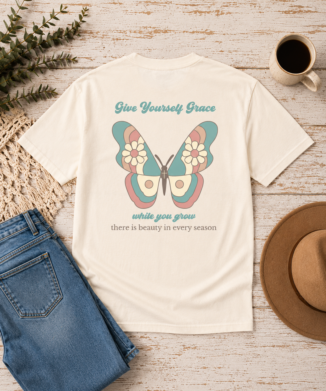 An Ivory comfort color graphic t-shirt displayed in a flat-lay arrangement on a rustic white-washed wooden background, styled with blue jeans, a brown hat, a coffee mug, and green foliage. The back of the t-shirt features a colorful vintage butterfly graphic with teal, pink, and blue tones, accompanied by the text 'Give Yourself Grace while you grow there is beauty in every season'.