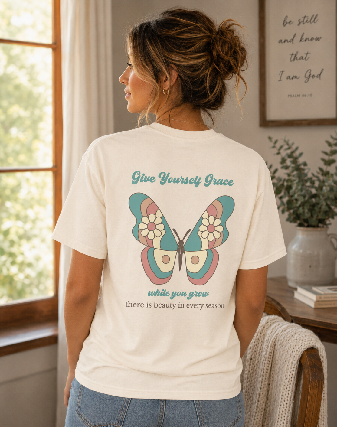 A woman shown from the back wearing an Ivory comfort colors vintage graphic t-shirt with a vintage butterfly design and the text 'Give Yourself Grace while you grow there is beauty in every season'. The setting is a warm, softly-lit room with a window, framed wall art, and a plant.
