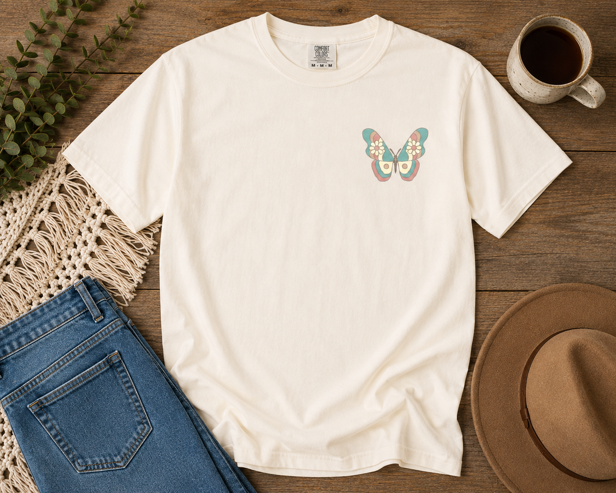 An Ivory comfort colors short-sleeve t-shirt with a colorful vintage butterfly graphic on the left chest, styled in a flat-lay arrangement on a wooden surface with blue jeans, a cup of coffee, a hat, and a woven mat.