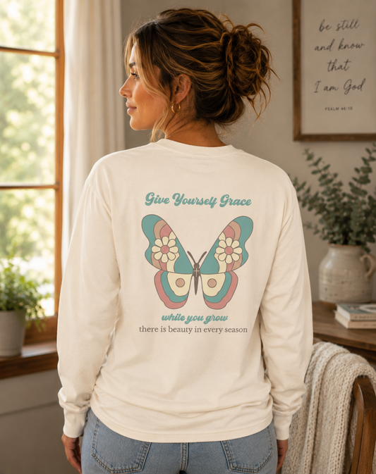 A woman shown from the back wearing an Ivory comfort color long-sleeve shirt with a colorful vintage butterfly graphic and the text 'Give Yourself Grace while you grow there is beauty in every season'. The setting is a cozy, sunlit room with plants and a framed quote on the wall.