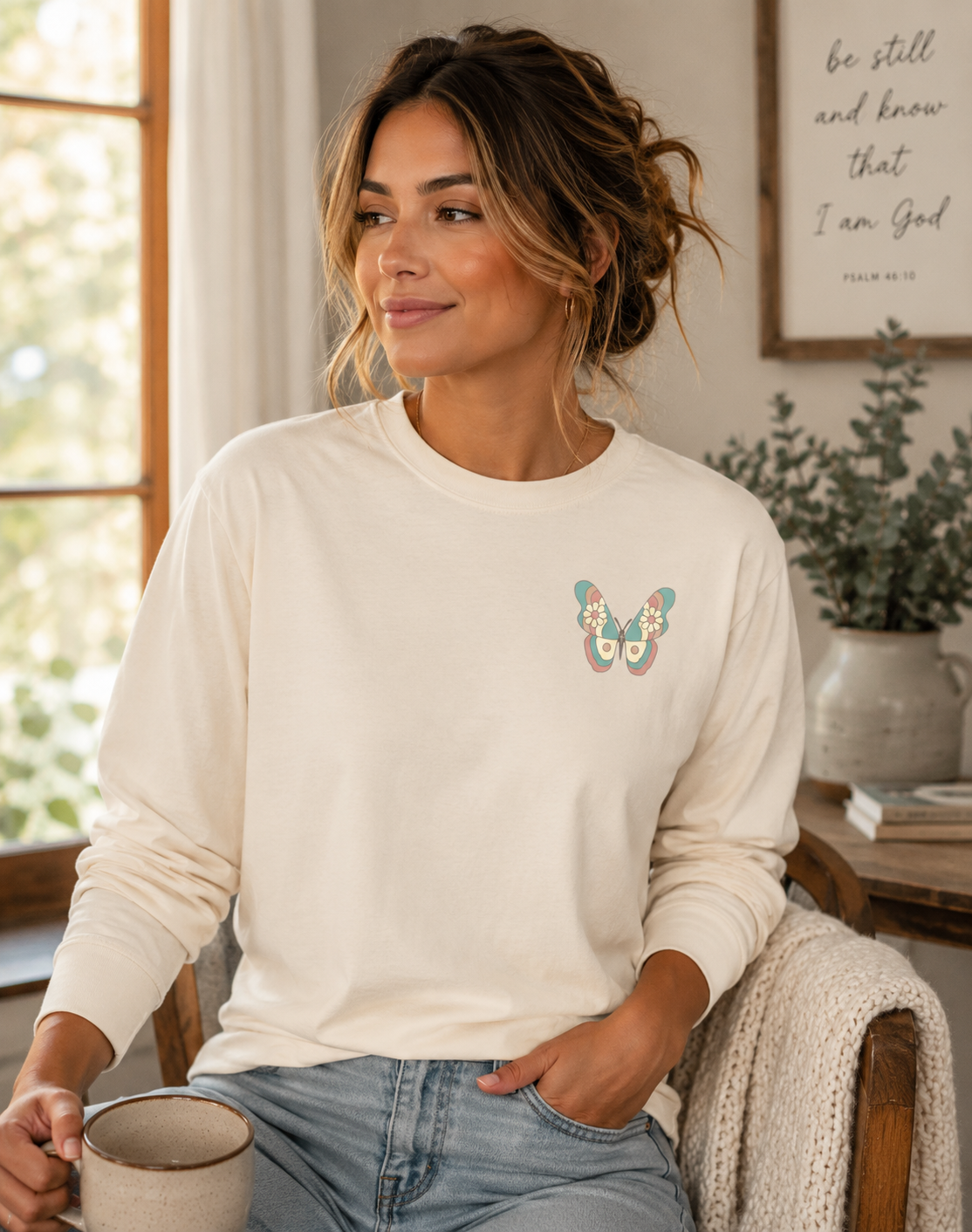 A woman sitting in a cozy room wearing an Ivory comfort colors long-sleeve t shirt with a colorful vintage butterfly graphic on the left chest, paired with light blue jeans.