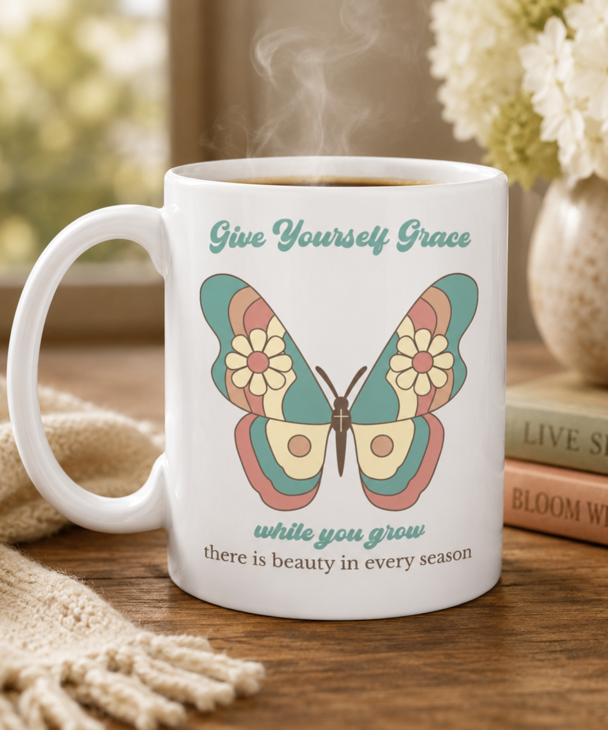 A white ceramic coffee mug with a colorful butterfly graphic and the text 'Give Yourself Grace while you grow there is beauty in every season', shown on a wooden table with a cozy background.