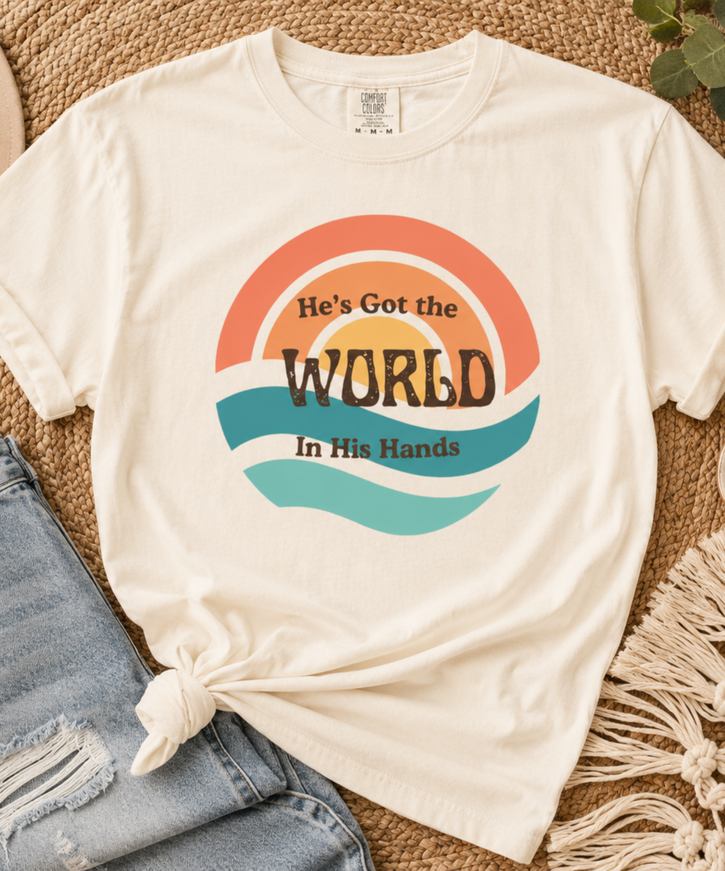 An Ivory comfort colors graphic t-shirt styled in a flat lay with a distressed denim jean leg tied at the hem, a beige straw hat, a white mug, and a macrame runner on a jute rug. The t-shirt features a retro sunset graphic with the text 'He's Got the WORLD In His Hands'.