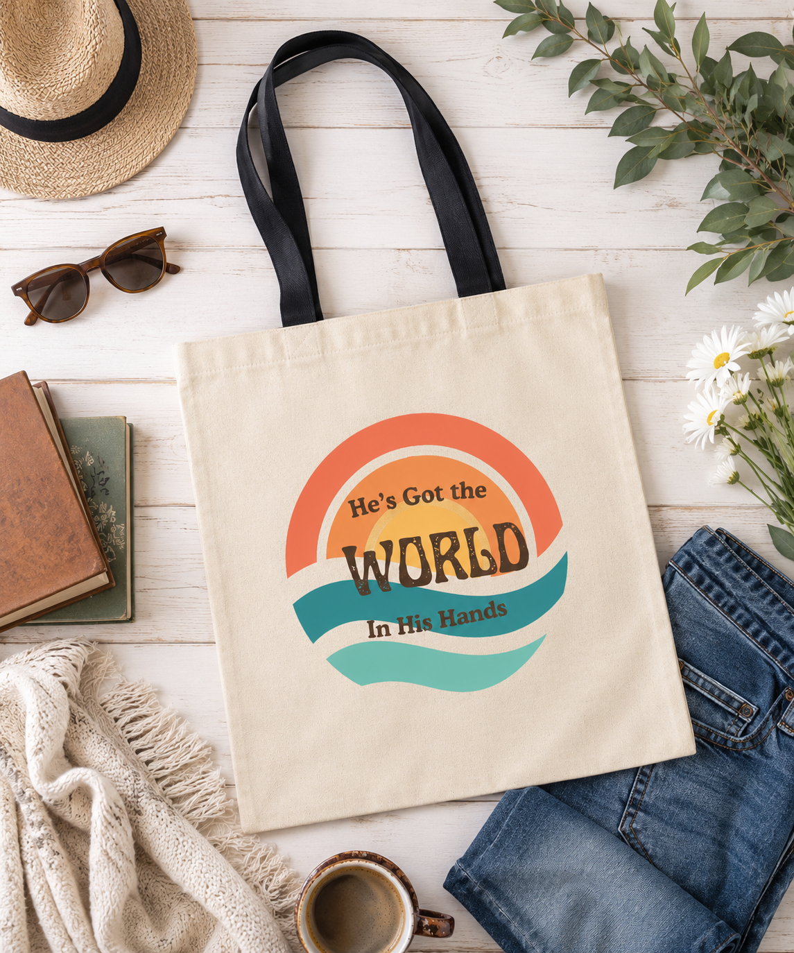 Flat lay of a natural cotton tote bag featuring the retro-inspired "He's Got the World in His Hands" design in warm coral, golden yellow, and teal tones, styled with books, denim, flowers, sunglasses, and cozy everyday accessories.