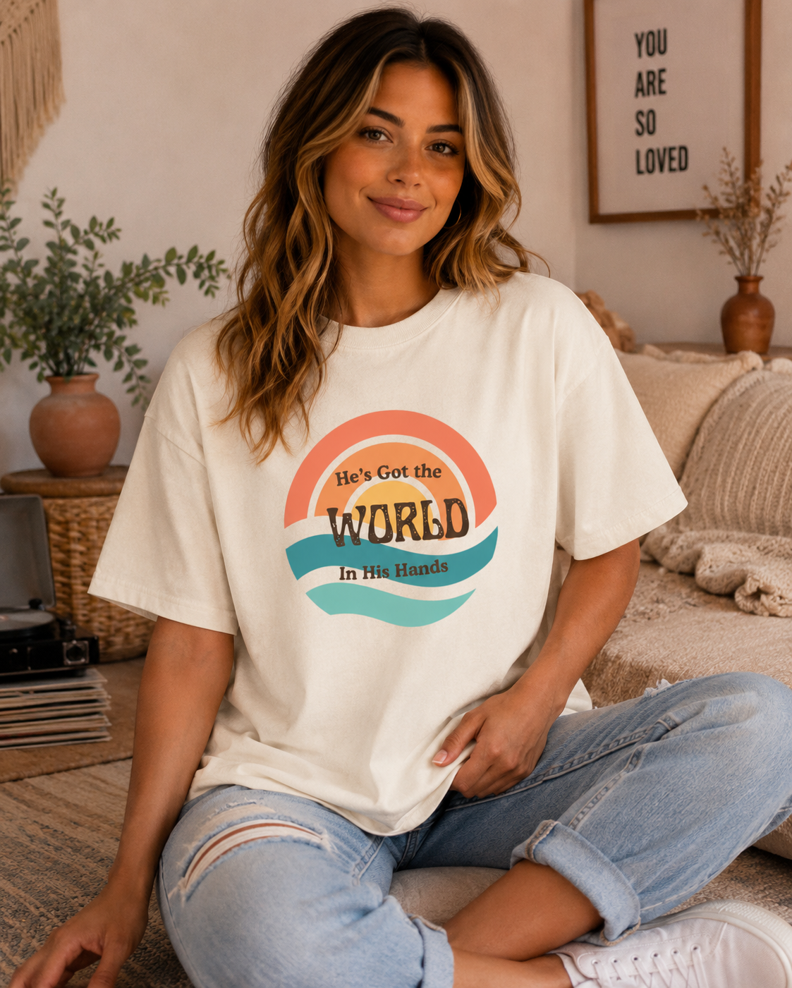 A woman sitting on a couch wearing a Ivory comfort colors graphic t-shirt with a retro rainbow design and the text 'He's Got the WORLD In His Hands'.