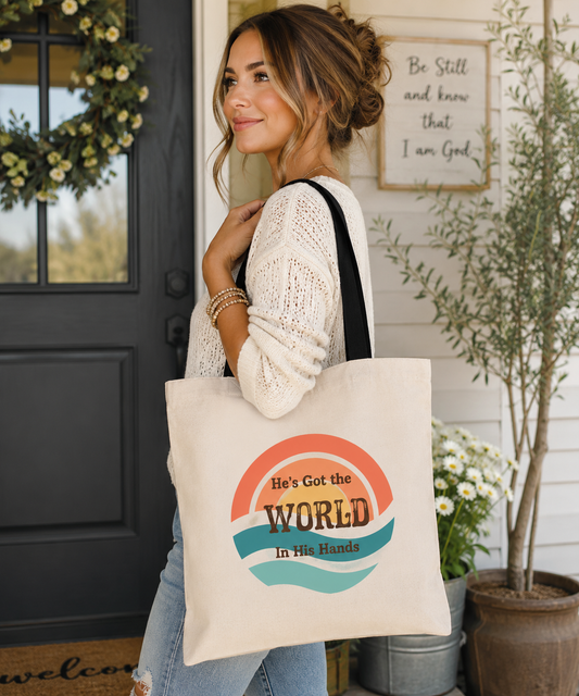 Woman carrying a natural cotton tote bag featuring the retro "He's Got the World in His Hands" design with vintage sunset colors, photographed in a warm and inviting porch setting.