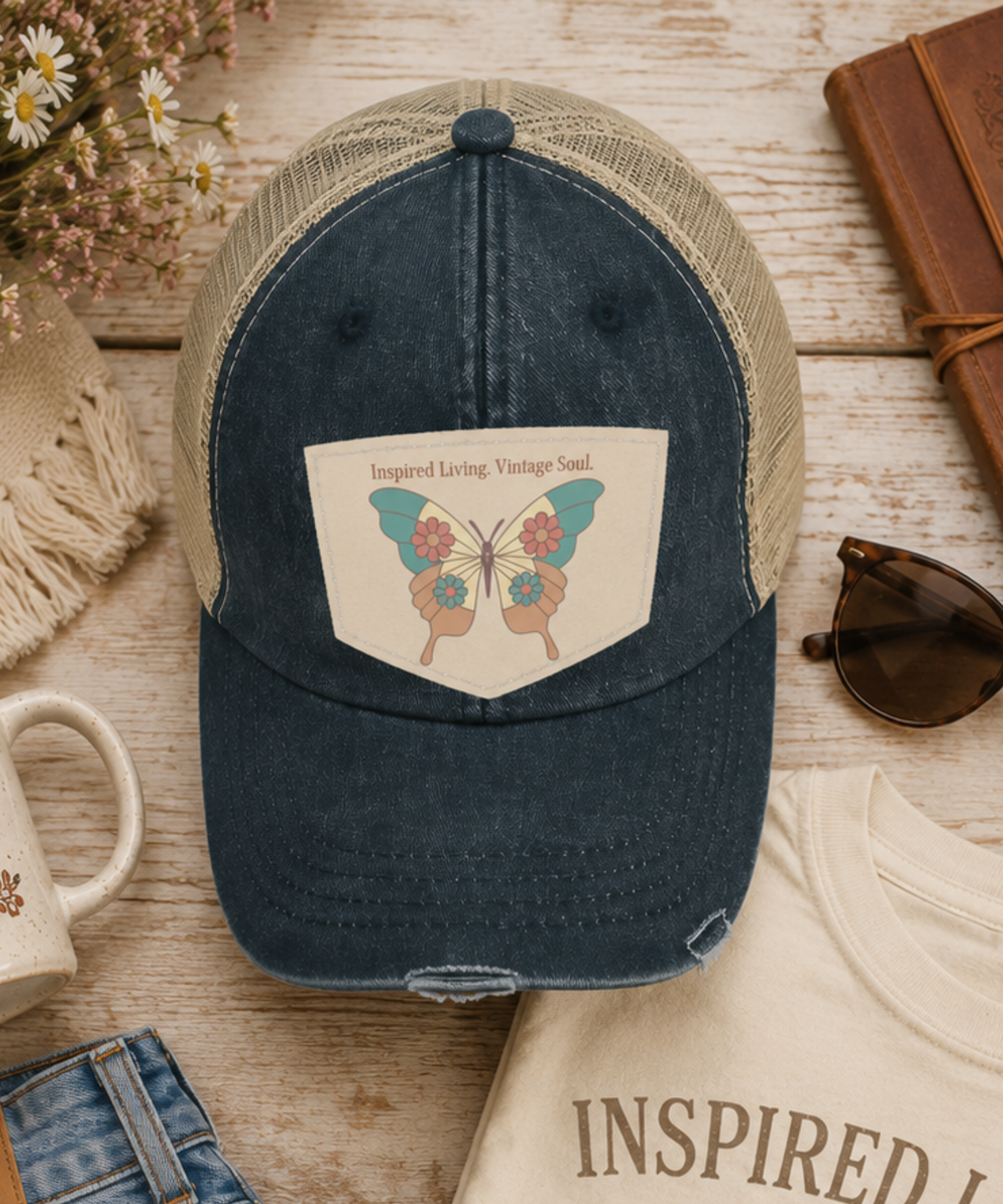 A distressed trucker cap with a navy washed front panel and beige mesh back, featuring a graphic patch on the front with butterfly wings and the text 'Inspired Living Vintage Soul'. The hat is styled in a flat-lay arrangement on a rustic wooden table with a mug, sunglasses, denim, and a cream t-shirt.