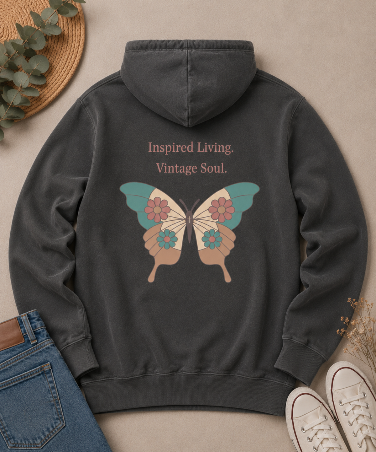 Flat lay of a Comfort Colors Pepper hoodie featuring the "Inspired Living. Vintage Soul." butterfly back design, styled with blue jeans, white canvas sneakers, eucalyptus leaves, and dried florals on a neutral beige background.
